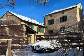 A stone building with a snow-covered roof and green shutters, a wooden fence, and a snow-covered ground. - Holiday Home 'Cottage des Pléiades' with Lake View and Wi-Fi (Les Angles)