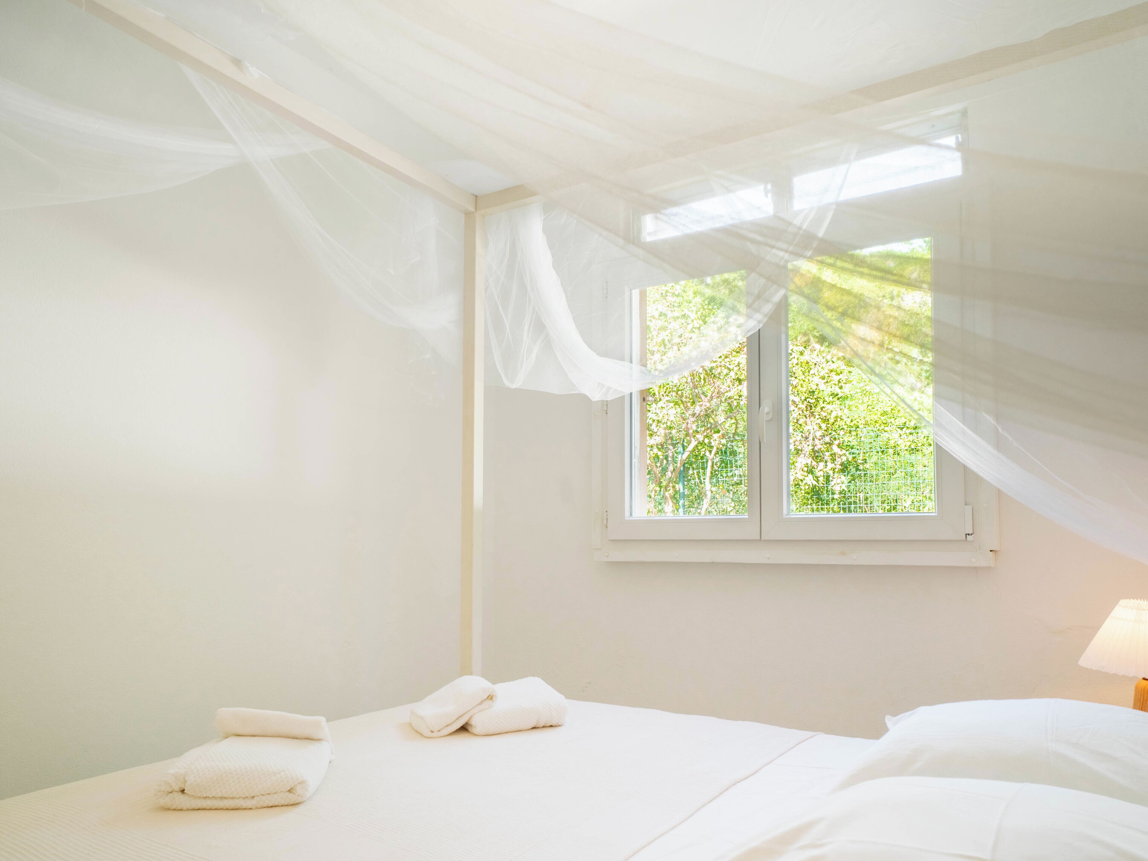 A bedroom with a bed, white curtains, and a window with a view of greenery.