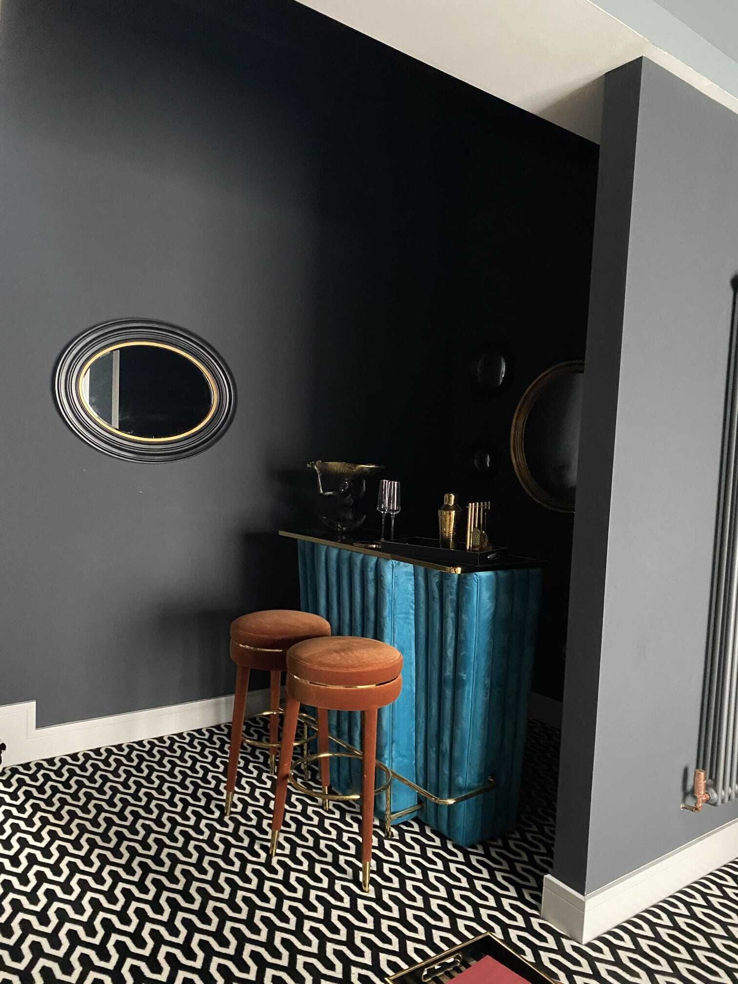 A room with a black and white patterned floor, two brown stools, a blue bar counter, and a round mirror on the wall.