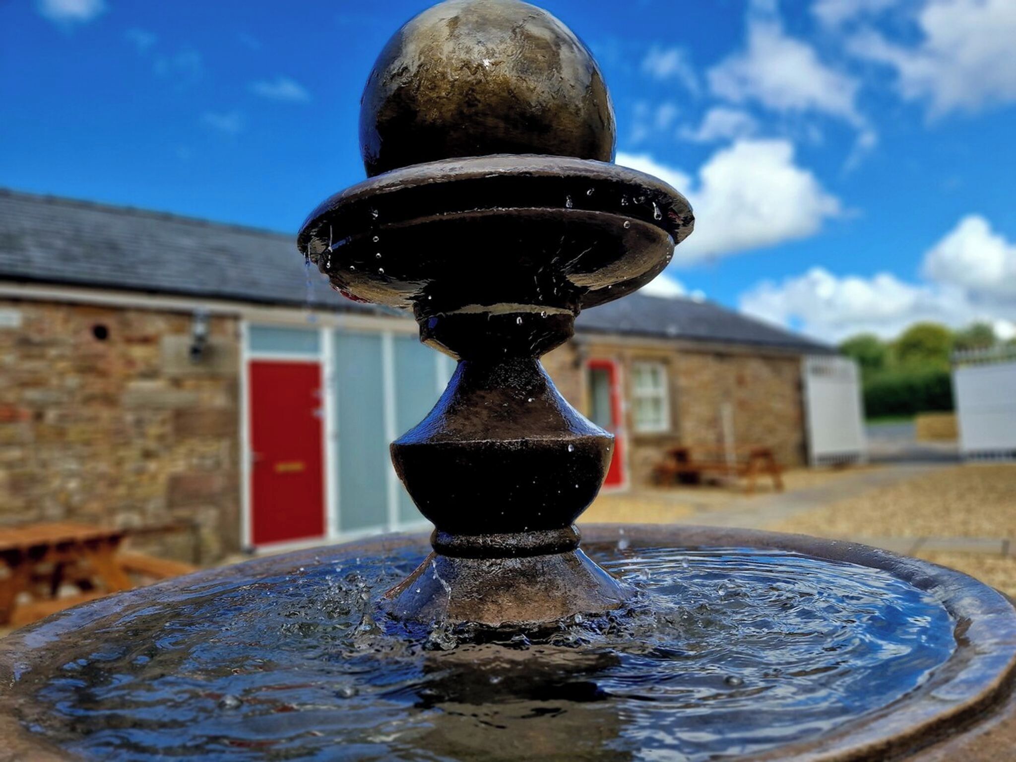 A fountain with a spherical top and a basin, set against a building with a red door and stone walls.