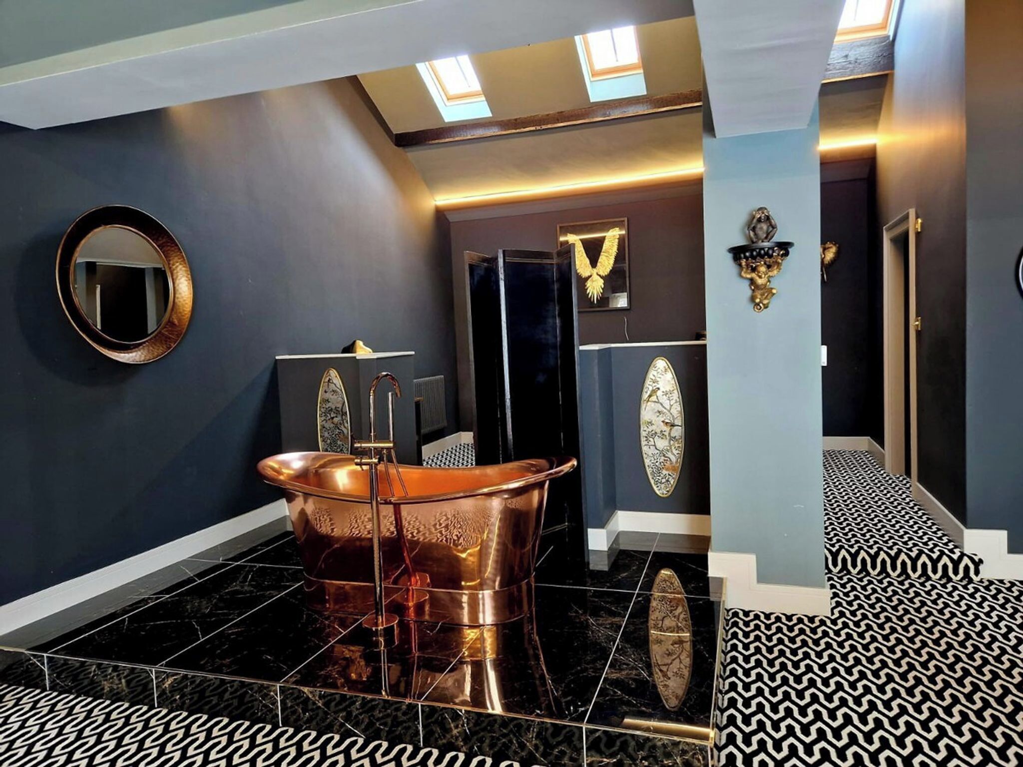 A bathroom with a copper bathtub, a round mirror, and decorative wall art.