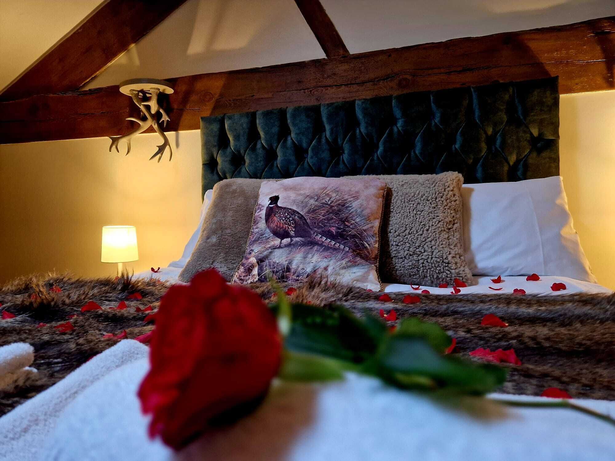 A bedroom with a tufted headboard, a bed with rose petals, a red rose, and a lamp.