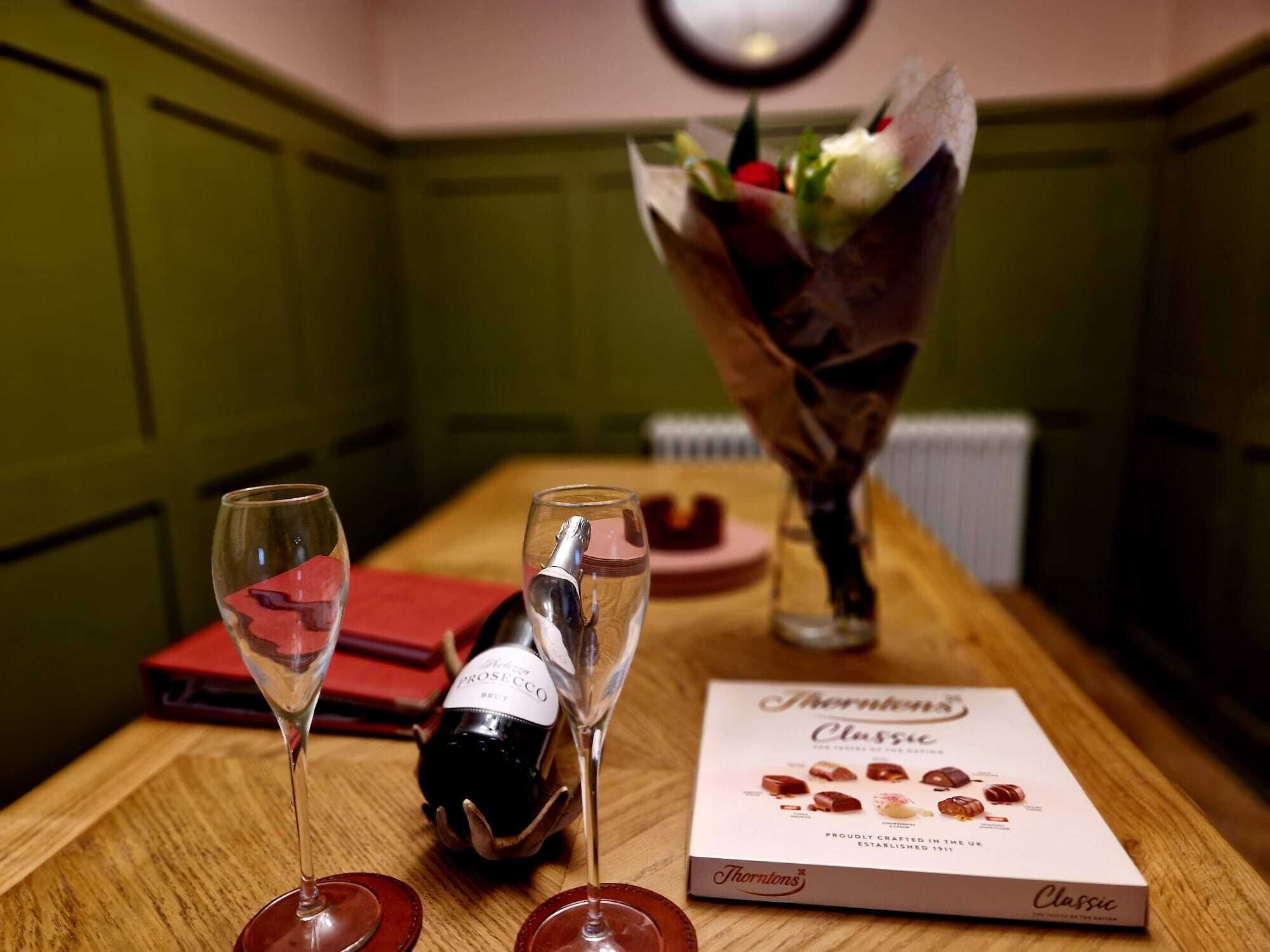 Una mesa con dos copas de vino, una botella, una caja de bombones y un jarrón con flores.