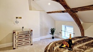 A loft bedroom with exposed wooden beams, a dresser, a potted plant, and a table with a bottle and grapes.
