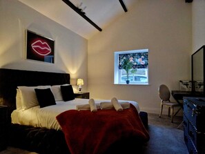 Un dormitorio con cama, una manta roja, una mesita de noche con lámpara, una ventana con vistas y un espejo.
