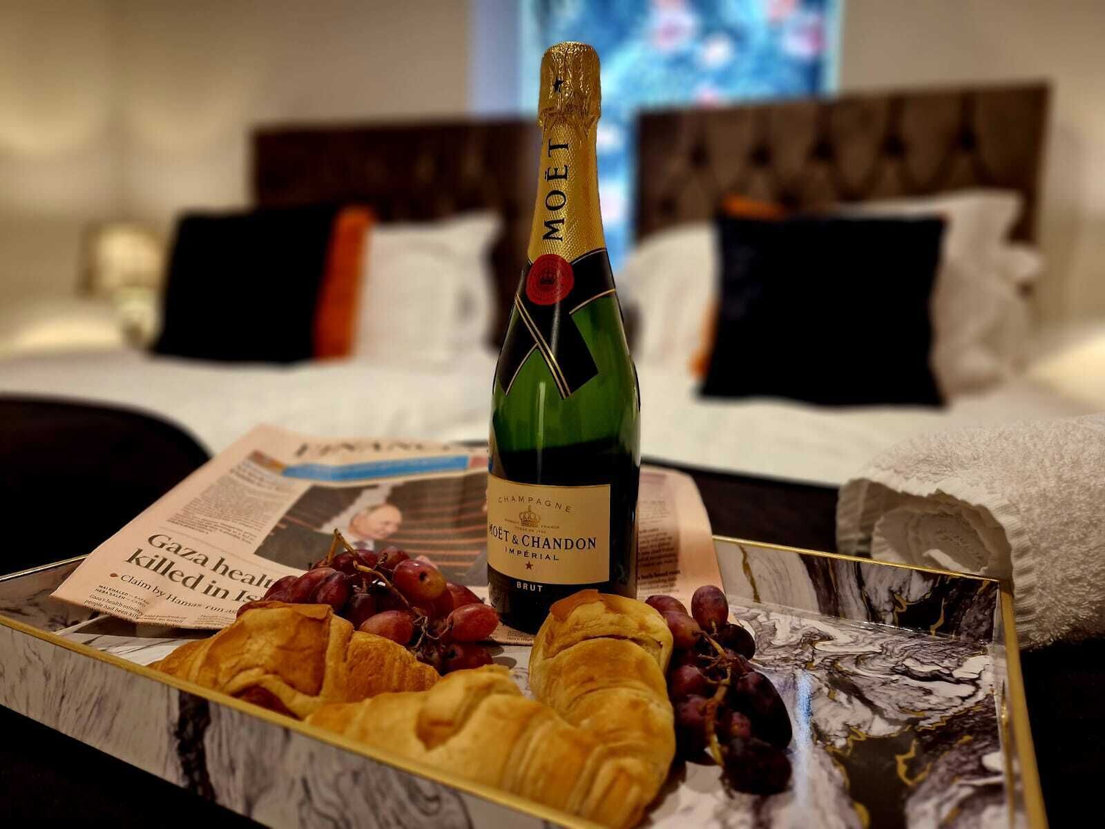 A bottle of Moët & Chandon champagne, a newspaper, and a tray with croissants and grapes on a bed with a headboard and pillows.