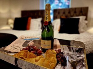 Una botella de champán Moët & Chandon, un periódico, y una bandeja con cruasanes y uvas sobre una cama con cabecera y almohadas.