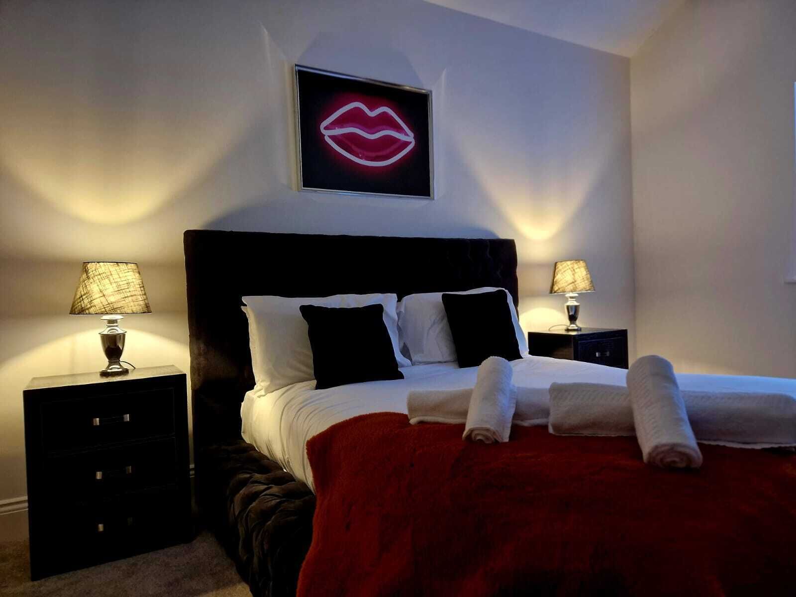 Ein ordentlich bezogenes Bett mit weißer und schwarzer Bettwäsche, zwei Nachttische mit Lampen, eine rote Tagesdecke und ein leuchtendes Schild mit Lippenbild auf der Wand.