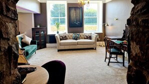 A cozy living room with a beige sofa, a green armchair, and a dining table with chairs. There is a chandelier hanging from the ceiling and a large window with a view of greenery outside. - The Hamilton luxury apartments-Lord William suite (Cabus)