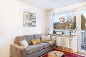 Un salon avec un canapé gris, des coussins à motifs, un radiateur, une fenêtre décorée de fleurs et une vue sur les bâtiments à l’extérieur.
