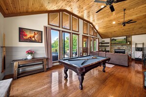 A pool table in a room with wooden flooring, a ceiling fan, and large windows.