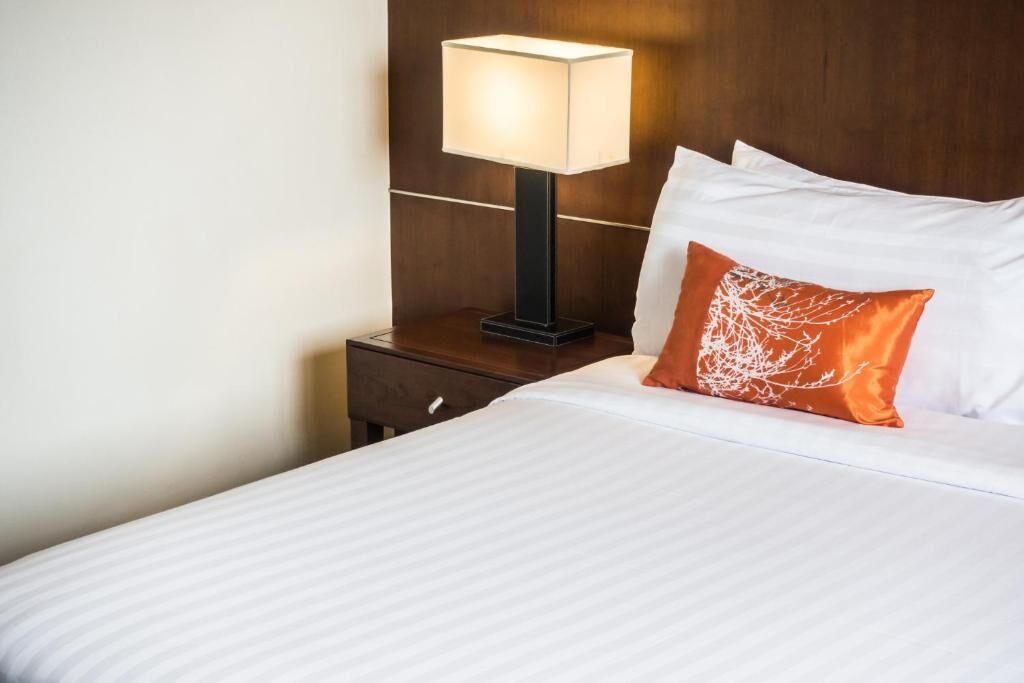 A neatly made bed with white linens and a single orange decorative pillow. A modern table lamp with a square shade is placed on a wooden nightstand beside the bed.