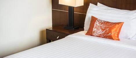 A neatly made bed with white linens and a single orange decorative pillow. A modern table lamp with a square shade is placed on a wooden nightstand beside the bed.