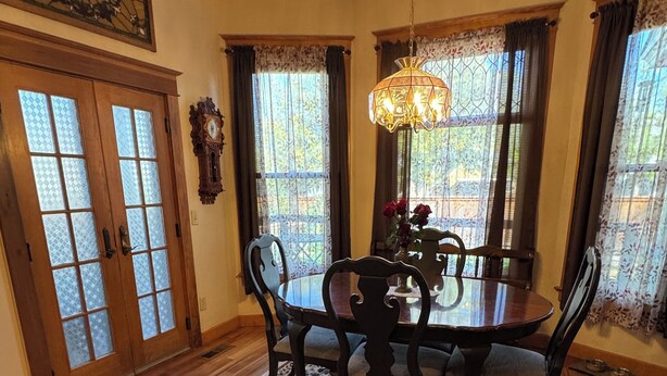 Ein Esszimmer mit Holztisch und -stühlen, einer Chandelierleuchtre und Blick durch große Fenster mit Vorhängen.