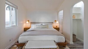 A bedroom with a large bed, two bedside tables with lamps, a bench at the foot of the bed, a window on the left, and an arched doorway to another room. - Stylish Bungalow Steps to Sand & Downtown MB (Manhattan Beach)