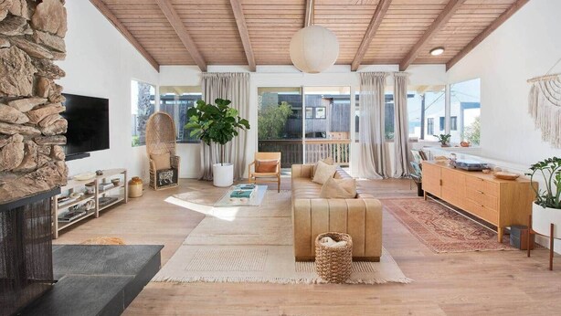 A spacious living room with a stone wall, wooden ceiling beams, a large TV, a sofa, a coffee table, and a dining area with a wooden cabinet.