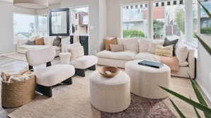 Ein modernes Wohnzimmer mit einem Ecksofa, runden Hocker und einem großen Fenster mit Blick ins Freie.
