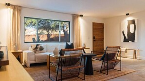 Un salon moderne comprenant un canapé, deux fauteuils, une table basse, une grande fenêtre donnant sur des arbres et une porte en bois.