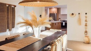Une cuisine moderne avec un coin repas, comprenant un grand luminaire suspendu en fibres tressées, un vase blanc orné de plumes d’herbe des pampas et une table à manger en bois.