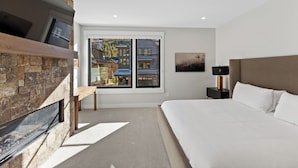 A bedroom with a stone fireplace, a large bed, a nightstand with a lamp, and a window with a view of buildings. - Ac39k4 (Keystone)