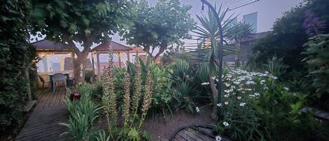 Um deck de madeira leva a um gazebo com luzes de corda, rodeado por vegetação exuberante e plantas floridas.
