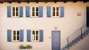 Un edificio de dos pisos con contraventanas azules, una puerta gris y una escalera que lleva al piso superior.