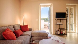 Una sala de estar con un sofá, cojines rojos, una mesita de centro, un televisor sobre un mueble y vistas a un balcón desde una puerta abierta.