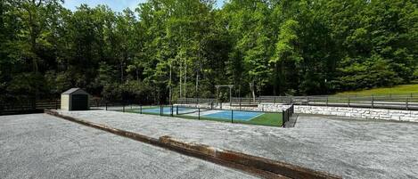 A concrete parking area with a small shed, a basketball court, and a fenced area with trees in the background.