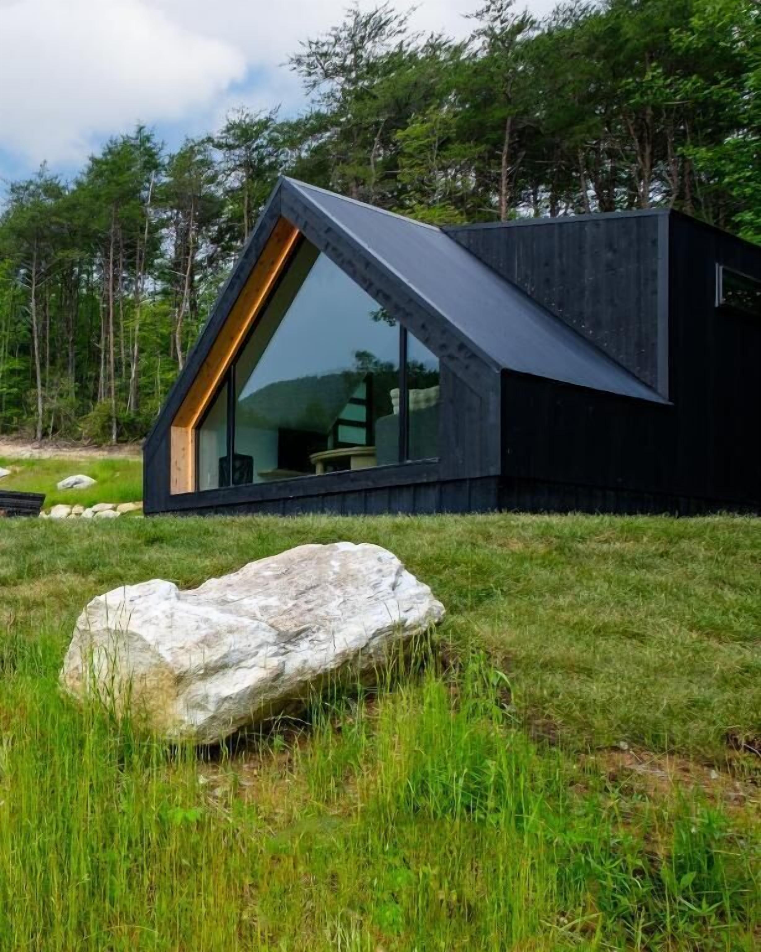 A modern cabin with a large glass window and a sloped roof, surrounded by a grassy area and a large rock.
