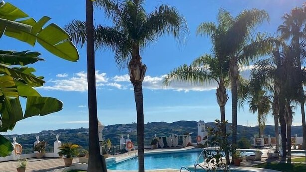 Zona de piscina con palmeras, cielo despejado y una cadena montañosa distante.
