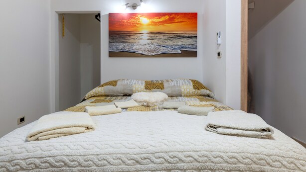 Un dormitorio con cama, almohadas y un cuadro colgado en la pared que muestra un atardecer sobre el océano.