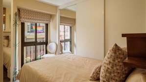 Une chambre à coucher comprenant un lit, une chaise, un miroir, une fenêtre donnant sur des immeubles et un luminaire au plafond.