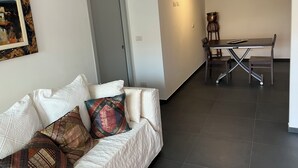 Una sala de estar moderna con un sofá, una mesa de comedor y una silla. Hay una puerta que da a otra habitación y un equipo de aire acondicionado en la pared.