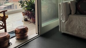 Una habitación con puerta corrediza de vidrio que da a un balcón con vistas a edificios y al atardecer. Hay un sofá blanco con cojines y una planta en maceta en el balcón.