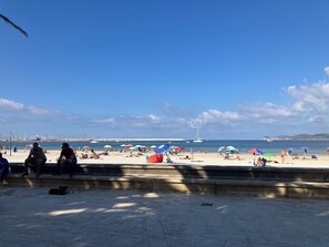 A beach with people relaxing under umbrellas, a dog on the pavement, and boats in the water.