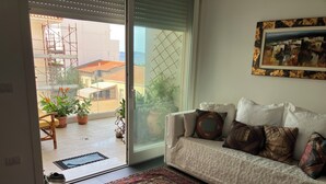 Una sala de estar con sofá, alfombra estampada y vistas a un balcón con plantas en macetas y una silla.