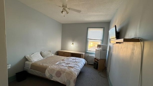 A bedroom with a bed, a desk, a radiator, and a ceiling fan. - City Limits Inn Hotel (Yorkton)