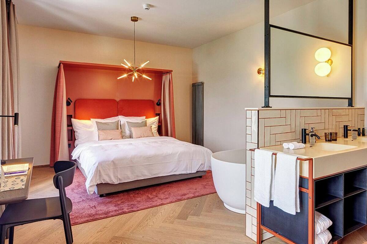 Moderni hotellihuone, jossa on sänky, työpöytä, tuoli, suuri peili, kylpyamme ja pesuallas.