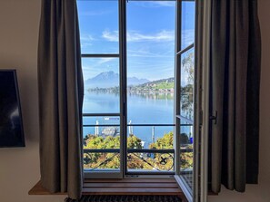 Et værelse med stort vindue med udsigt over en sø, en balkon med gelænder og trægulv.