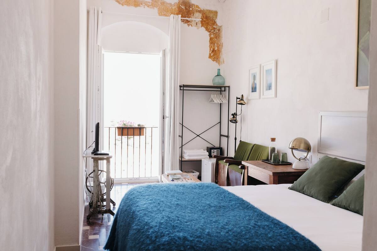A bedroom with a bed, a desk, a chair, a radiator, a window, and a balcony.
