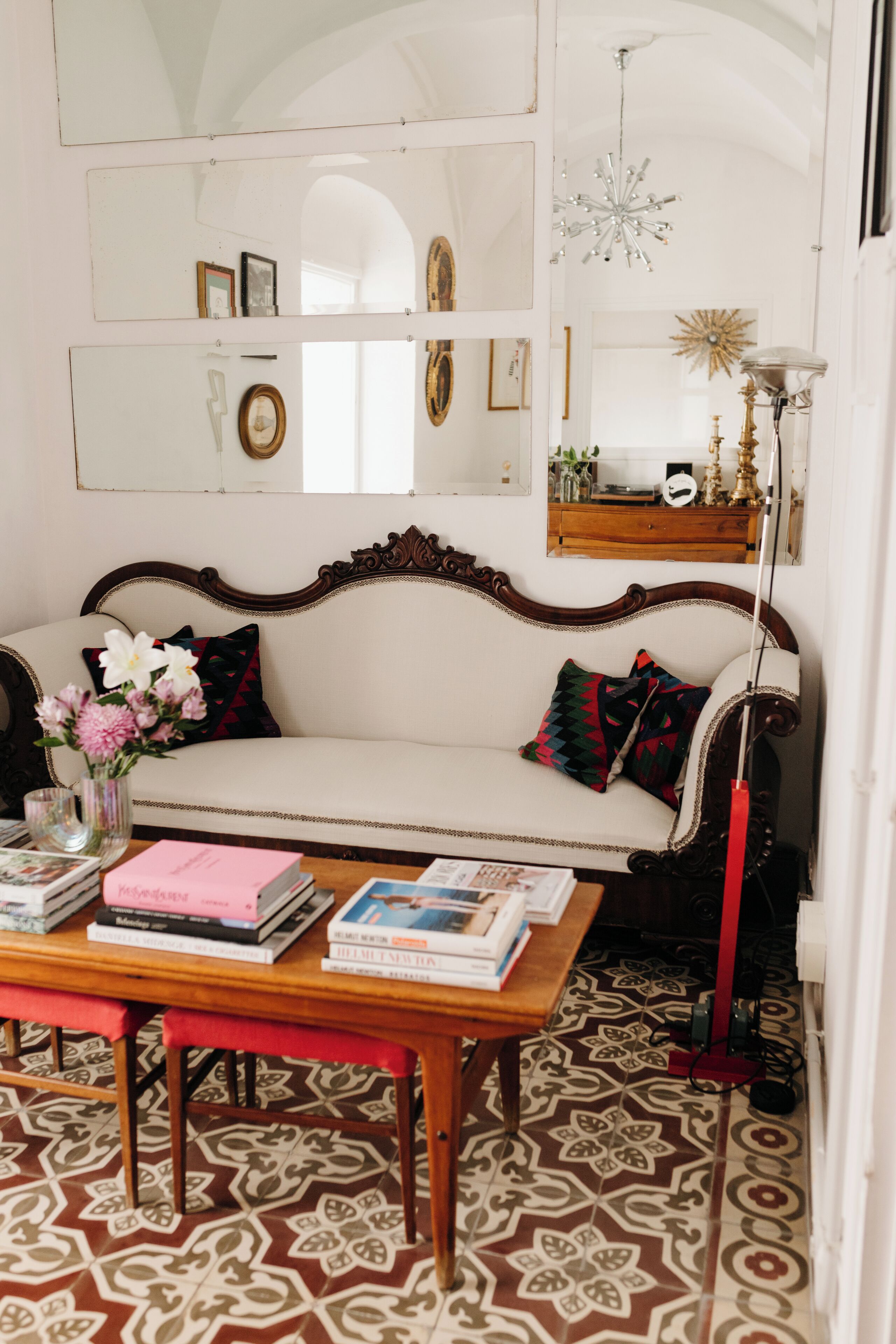 A cozy living room with a white sofa, a wooden coffee table with books, a patterned floor, and a chandelier.