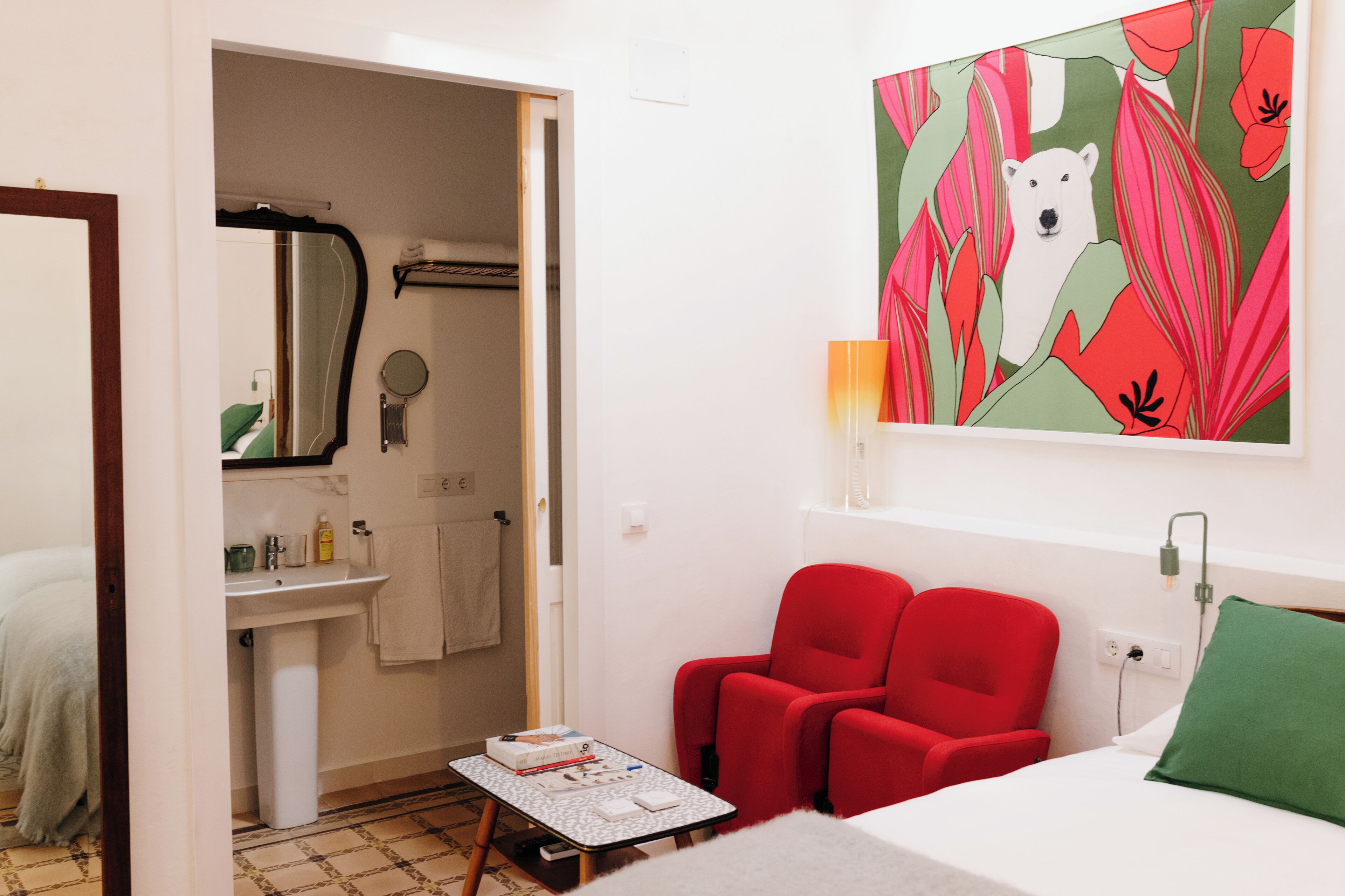 A hotel room with a bed, a red armchair, a small table with items, a bathroom with a sink and mirror, and a painting on the wall.