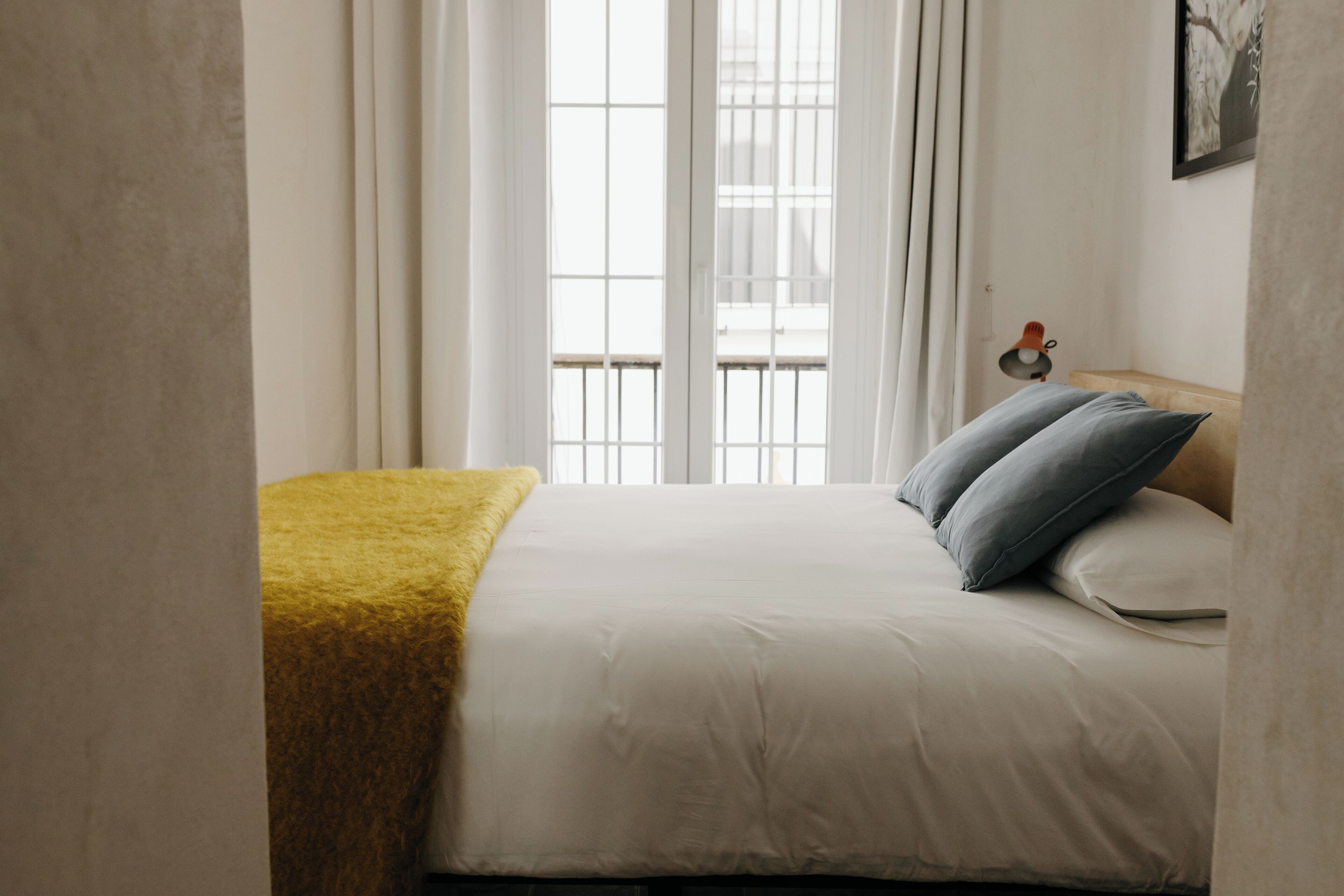 A neatly made bed with a yellow throw blanket and two pillows, a window with sheer curtains, and a small table with a lamp beside the bed.