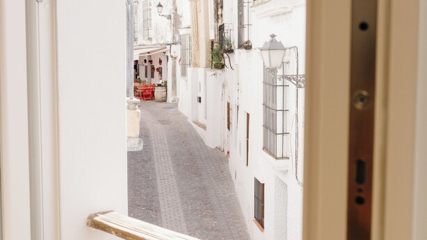 A narrow street with buildings on either side, a red table and chairs in the distance, and a metal railing in the foreground. - Botánico Casa Rural (Arcos de la Frontera)