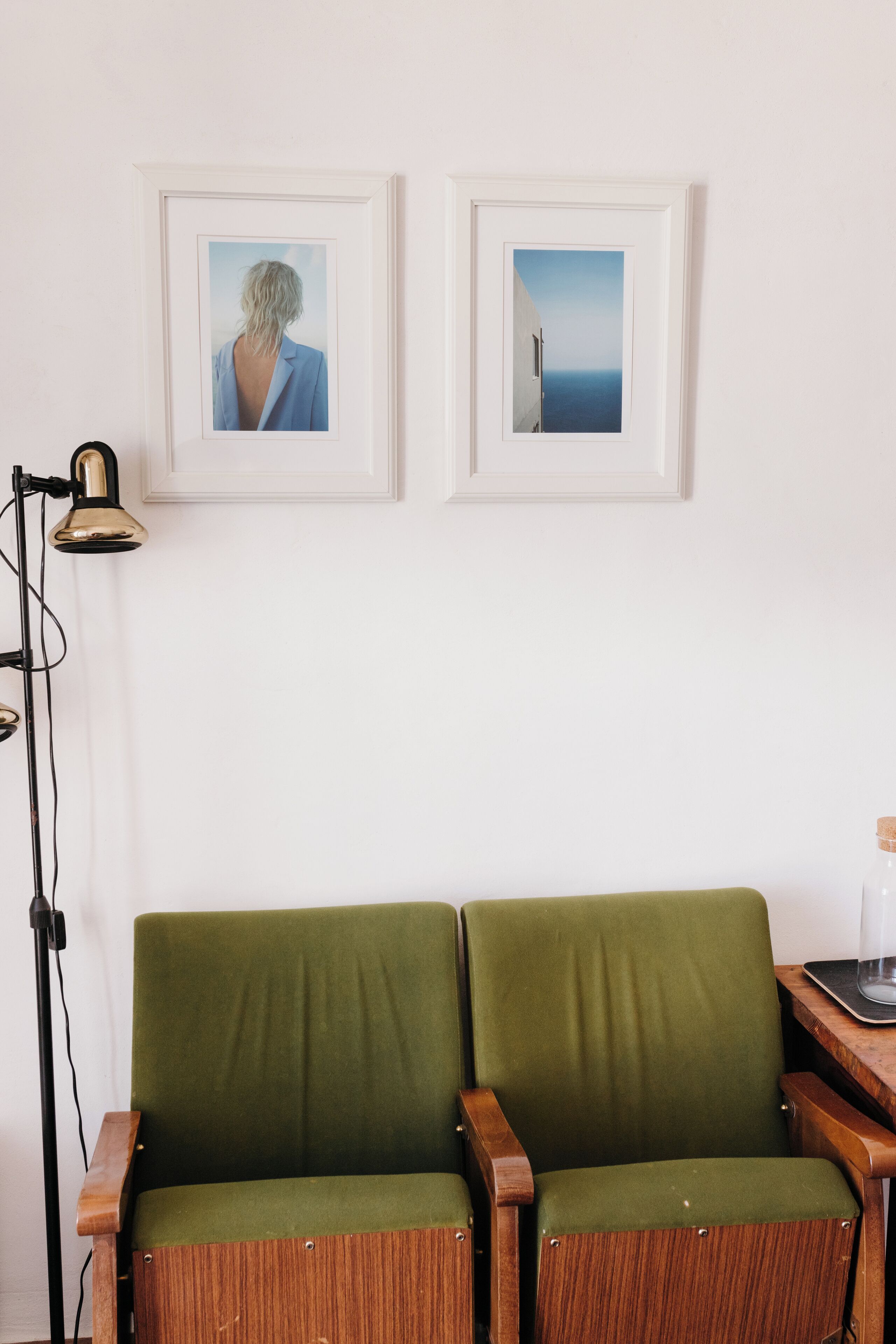 A room with two green upholstered chairs, a floor lamp, and two framed pictures on the wall.