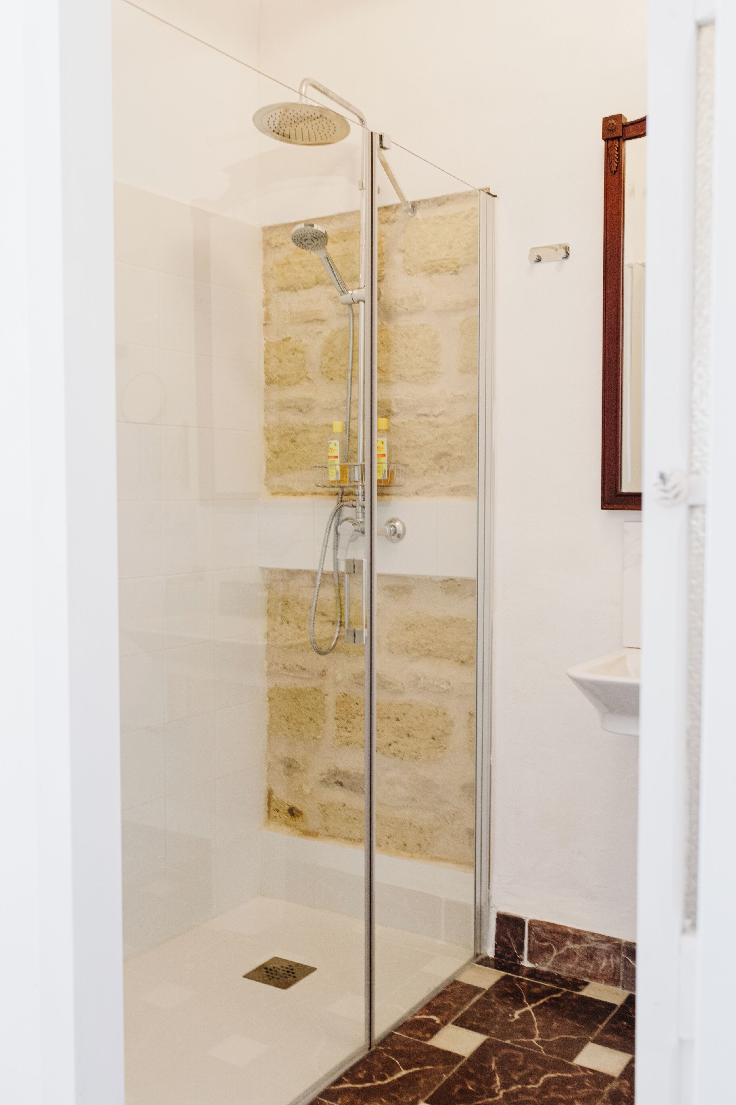 A glass-enclosed shower with a stone wall backdrop, a white sink, and a mirror.