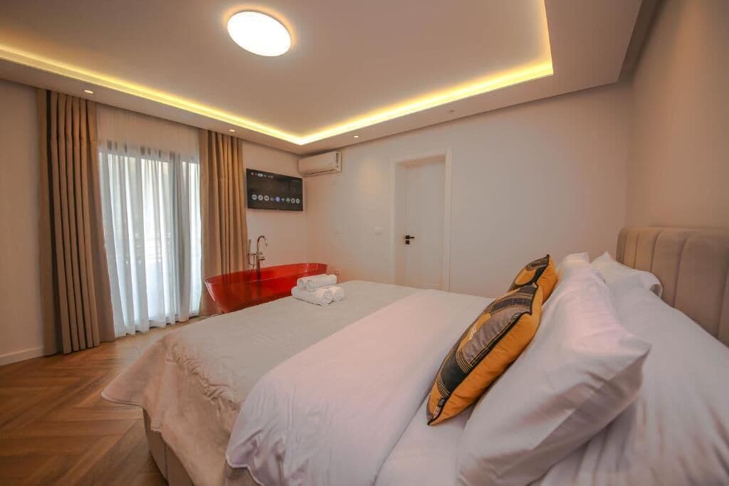 A hotel room with a bed, a red bathtub, a flat-screen TV, and a ceiling light.