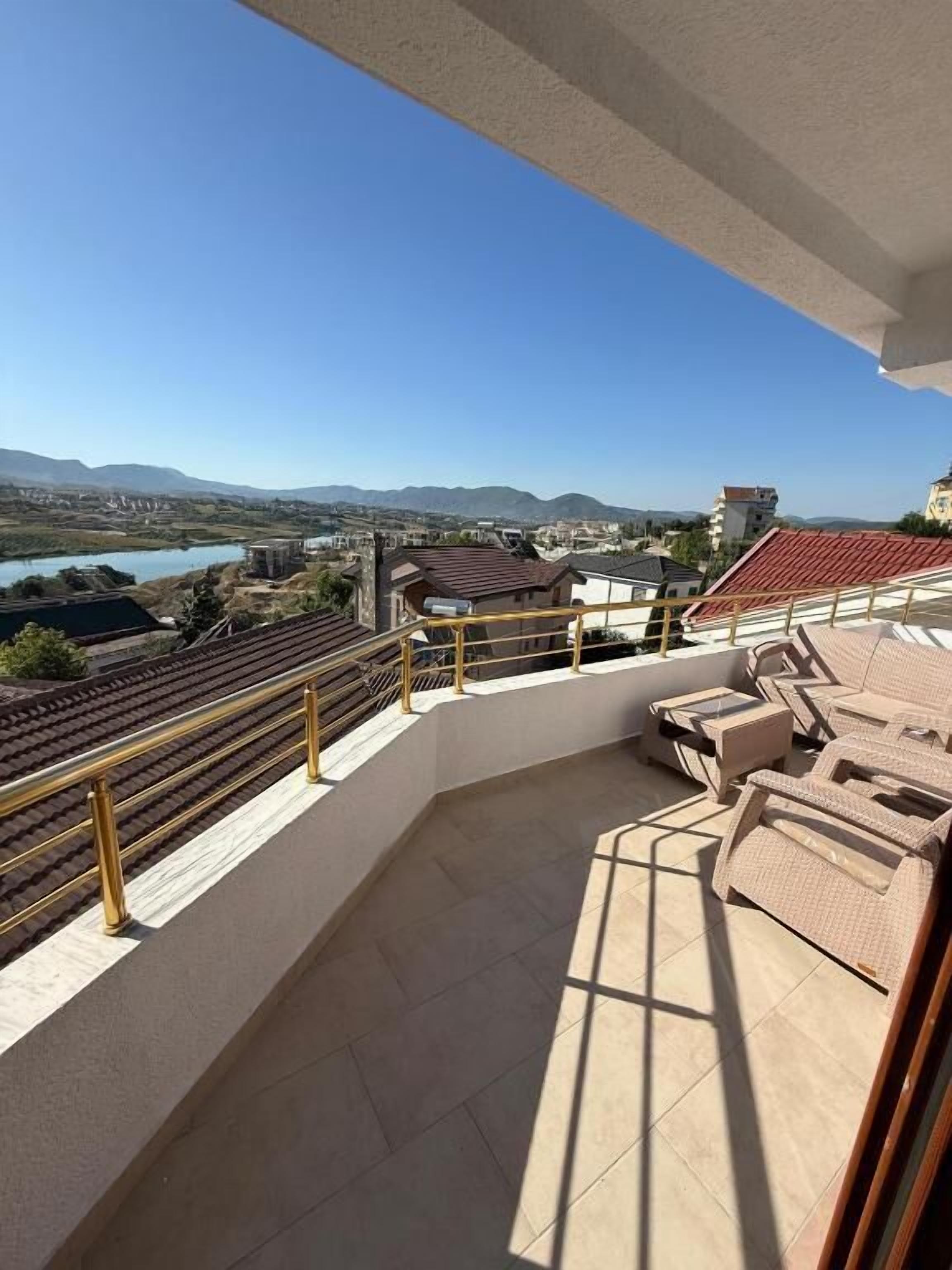 Un balcón con vista a un lago y montañas, muebles de exterior y suelo de baldosas.