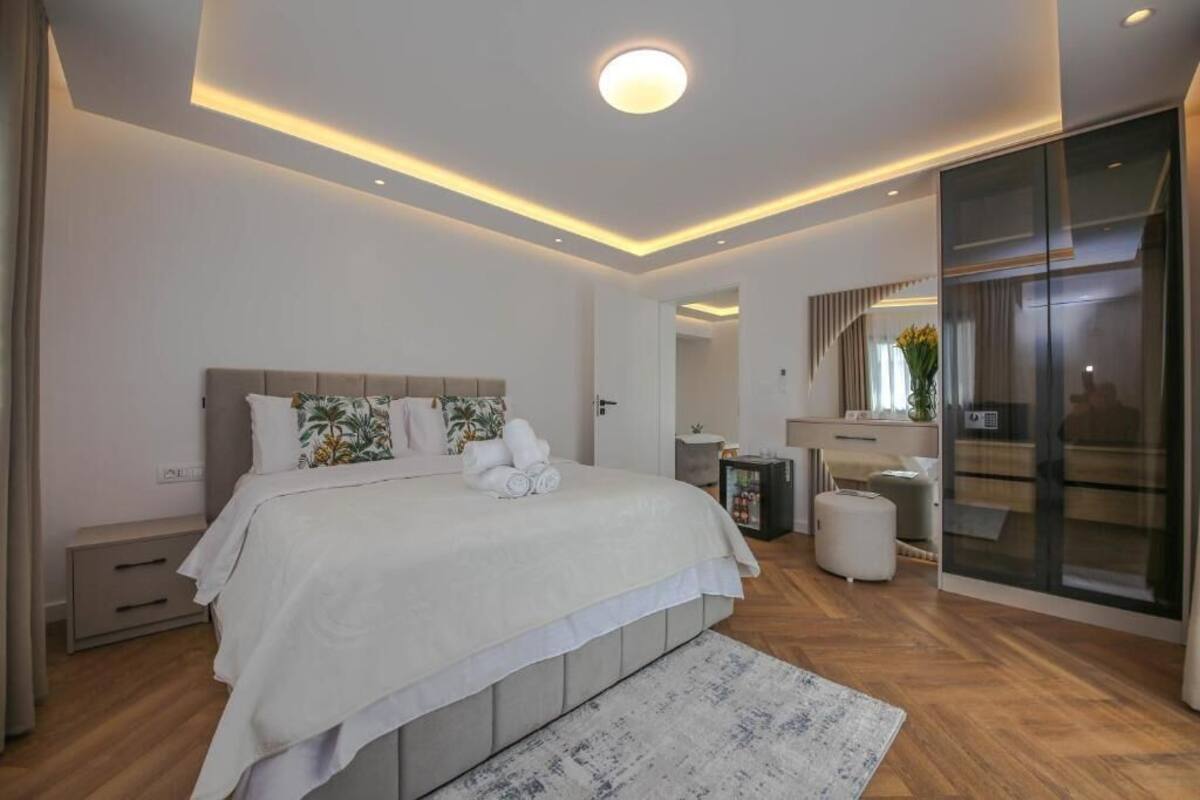 A modern bedroom with a large bed, bedside tables, a nightstand, a dresser, and a glass-enclosed walk-in closet.