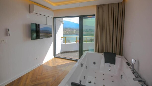 A modern bathroom with a large bathtub, a television, and a sliding glass door leading to a balcony with a view. - Murati Lake View Hotel (Farkë)